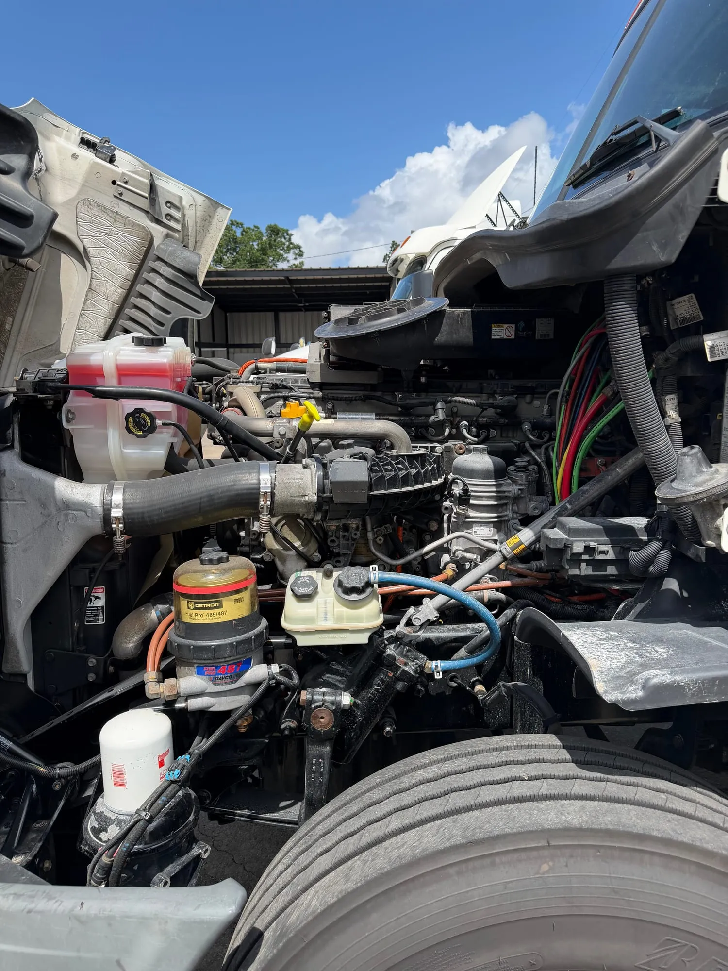 Hood tilted forward on a Detroit-powered semi truck with the engine bay open for service — coolant surge tank, charge-air piping, radiator hose and a Detroit fuel filter all in reach.